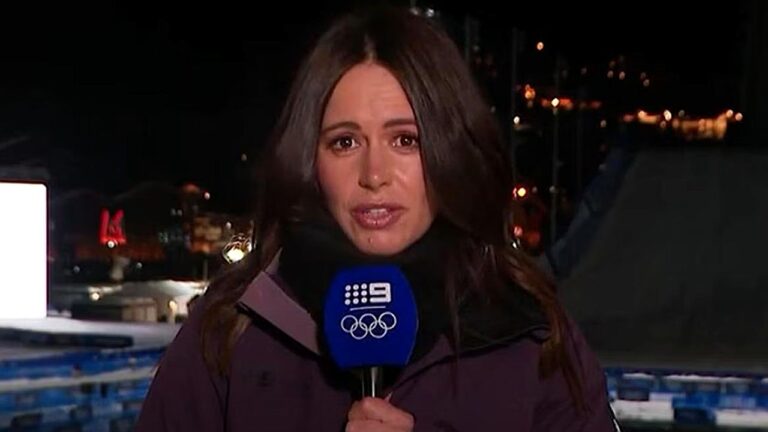 Repórter aparece em transmissão ao vivo durante a noite, segurando microfone com logotipo da emissora e símbolo olímpico. Ela veste casaco escuro e olha diretamente para a câmera com expressão séria, enquanto luzes da cidade e estruturas iluminadas desfocadas aparecem ao fundo, indicando cobertura jornalística em ambiente externo.