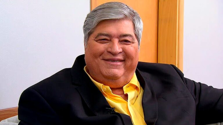 Homem de cabelos grisalhos sorri para a câmera enquanto está sentado em ambiente interno. Ele veste camisa amarela e blazer preto, em cenário que sugere entrevista ou participação em programa de televisão.