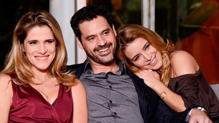 Três pessoas estão sentadas juntas em um sofá, sorrindo e aparentando estar em um momento descontraído. No centro, um homem de camisa cinza e blazer escuro sorri amplamente. À esquerda, uma mulher de vestido vermelho também sorri para a frente. À direita, outra mulher, com vestido escuro, apoia a cabeça no ombro do homem, demonstrando carinho. O ambiente parece interno e ac