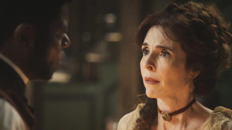 Mulher com expressão emocionada encara um homem em cena ambientada em época antiga. Ela tem cabelos castanhos presos em penteado elaborado, usa vestido em tom claro e gargantilha escura com detalhe central. A iluminação é baixa e quente, sugerindo ambiente interno à noite, possivelmente iluminado por velas, reforçando o clima dramático do diálogo.