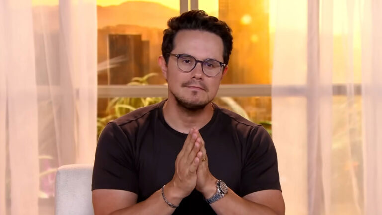 Deive Leonardo de camiseta preta e óculos, com mãos juntas em sinal de oração ou agradecimento, em ambiente com luz quente e janelas ao fundo