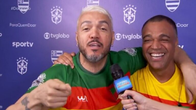 Dois homens aparecem abraçados durante uma entrevista em frente a um painel com logotipos da CBF e das emissoras TV Globo e SporTV. Ambos usam camisetas nas cores verde, amarelo e vermelho. O homem à esquerda, com cabelo curto e grisalho e barba aparada, fala animadamente enquanto gesticula; o da direita sorri amplamente