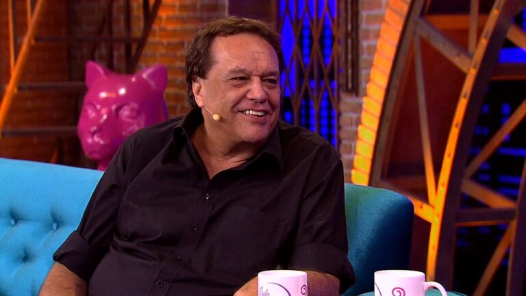 Homem de meia-idade está sentado em um sofá azul em um estúdio de TV. Ele veste camisa preta e usa um microfone de lapela no rosto. Está sorrindo e olhando para alguém ao lado, em clima descontraído. Ao fundo há um cenário com parede de tijolos, iluminação colorida em azul e laranja e um objeto decorativo rosa em formato de porco. Duas canecas estão apoiadas à frente, sugerindo um programa de entrevistas ou talk show.