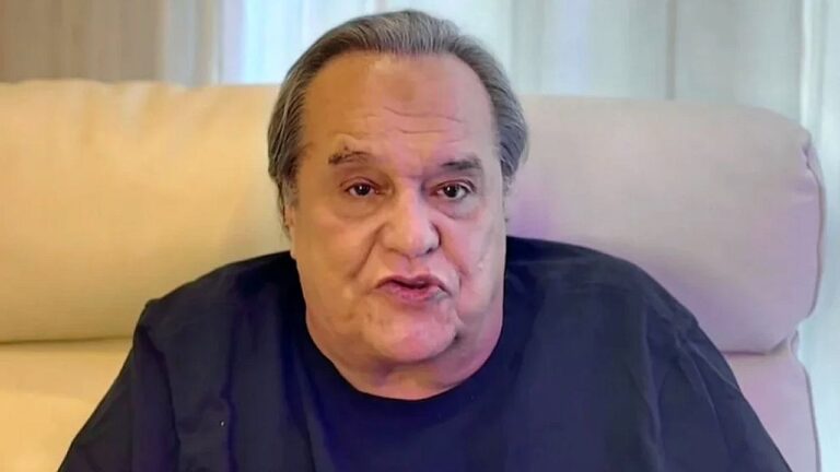 Homem mais velho, com cabelo grisalho penteado para trás, está sentado em uma poltrona clara em ambiente interno. Ele veste camiseta preta e parece estar falando, com expressão séria e concentrada. O fundo tem cortinas claras e iluminação suave, sugerindo uma gravação ou entrevista em casa ou escritório.