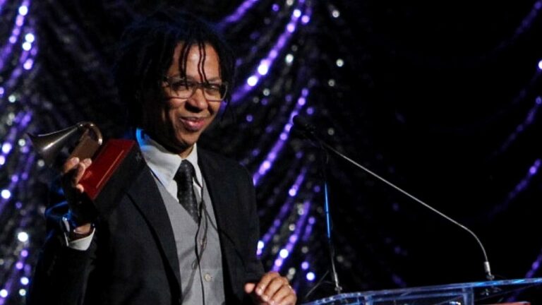 Pessoa com cabelos em dreadlocks e óculos segura um troféu em formato de gramofone enquanto discursa em palco iluminado. Vestindo terno escuro com colete e gravata, ela está diante de um púlpito com microfone, em cerimônia de premiação com fundo decorado por luzes roxas.