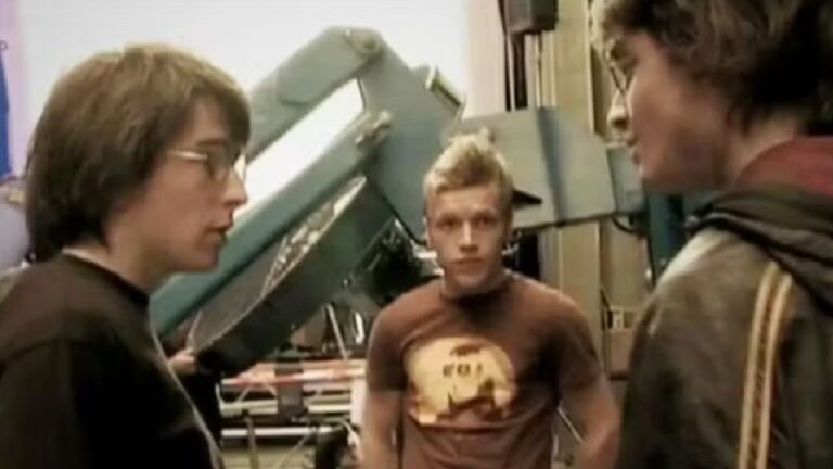 Três jovens estão em um ambiente que parece ser um estúdio ou sala com equipamentos grandes ao fundo. Dois deles usam óculos e conversam enquanto o rapaz ao centro, de camiseta marrom com estampa, observa com expressão séria.