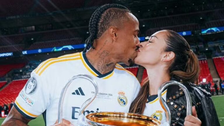 Duas pessoas se beijam em um estádio de futebol, ambas vestindo camisetas brancas do mesmo time com detalhes em dourado, enquanto seguram um grande troféu metálico de alças curvas, com gramado e arquibancadas ao fundo iluminadas por refletores. A pessoa à esquerda tem pele morena, cabelo trançado preso para trás, tatuagens visíveis no pescoço e usa uniforme esportivo, segurando uma das alças do troféu