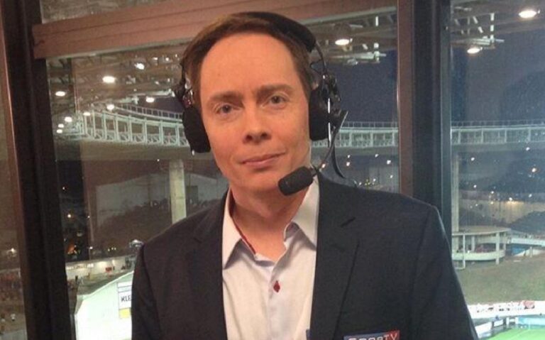 Narrador esportivo com headset e paletó do SporTV em cabine de transmissão com estádio iluminado ao fundo