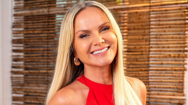 Mulher loira sorri para a câmera em um ambiente interno com persianas de madeira ao fundo. Ela usa vestido vermelho de gola alta e brincos dourados arredondados. A iluminação é quente e destaca sua expressão alegre e amigável.