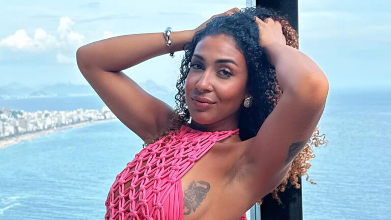 Emelly Souza posa com as mãos nos cabelos cacheados vestindo top de crochê rosa em sacada com vista panorâmica para o mar e orla da praia ao fundo