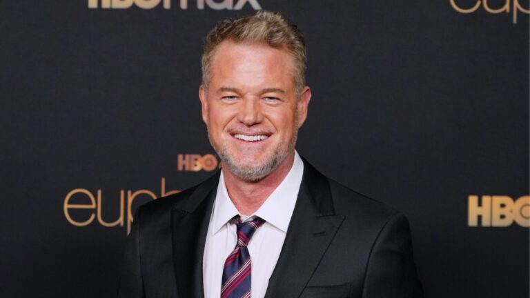 Eric Dane, um homem de cabelos grisalhos curtos e barba por fazer, sorri para a câmera em evento de tapete vermelho, vestindo terno preto, camisa branca e gravata listrada; ao fundo, painel com logotipos da HBO e da série Euphoria.