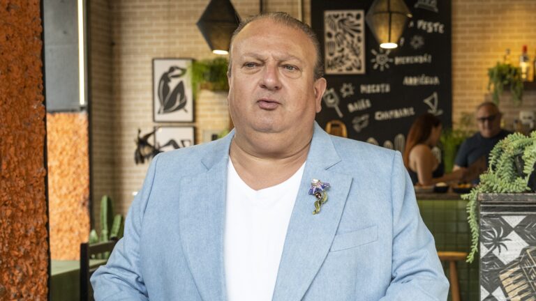 Erick Jacquin, com expressão séria, veste blazer azul-claro sobre camiseta branca e usa broche colorido na lapela, em ambiente interno de um restaurante do programa Pesadelo na Cozinha, da Band.