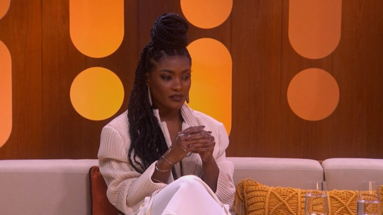 Mulher negra aparece sentada em um sofá de estúdio, em plano médio, com postura ereta e expressão séria e contemplativa. Ela tem cabelo longo em tranças, preso no topo da cabeça em um coque alto e volumoso, com algumas tranças caindo sobre o ombro. Usa maquiagem discreta e elegante, com destaque para os olhos.