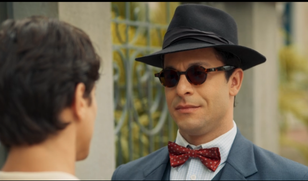 Um homem vestido com terno azul, camisa clara e gravata borboleta vermelha com bolinhas encara outro homem à sua frente. Ele usa chapéu preto e óculos escuros redondos, mantendo expressão séria e controlada. A cena acontece ao ar livre, diante de um portão ou grade metálica, sugerindo um encontro formal ou uma conversa tensa.