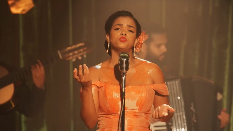 Mulher negra aparece em primeiro plano cantando em um palco, posicionada de frente para um microfone antigo de pedestal em estilo retrô, com expressão intensa e emotiva, boca aberta em canto e sobrancelhas levemente franzidas. Ela usa vestido longo em tom alaranjado com brilho, de alças caídas nos ombros, tecido ajustado ao corpo e superfície levemente refletiva. O cabelo está preso para trás em penteado elegante, adornado com uma flor laranja na lateral da cabeça, e ela usa brincos longos delicados.