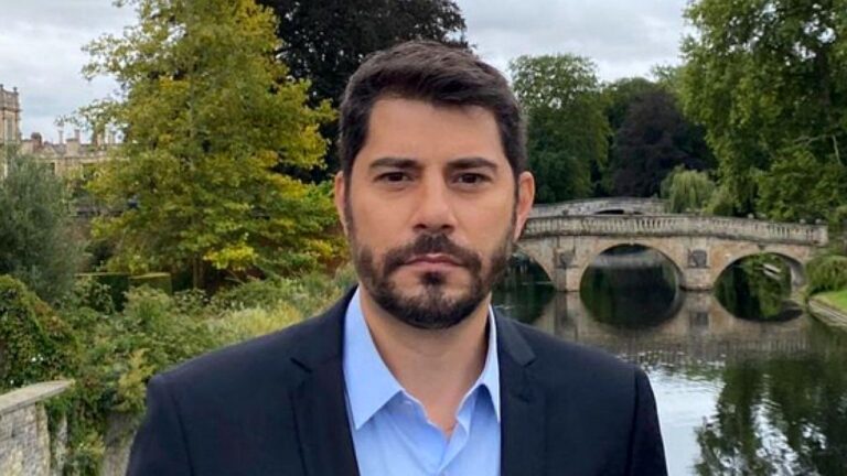 Homem de barba e terno escuro posa ao ar livre diante de rio e ponte de pedra ao fundo. Com expressão séria, ele olha diretamente para a câmera em cenário com árvores e construções históricas, sugerindo gravação externa ou reportagem em local turístico.