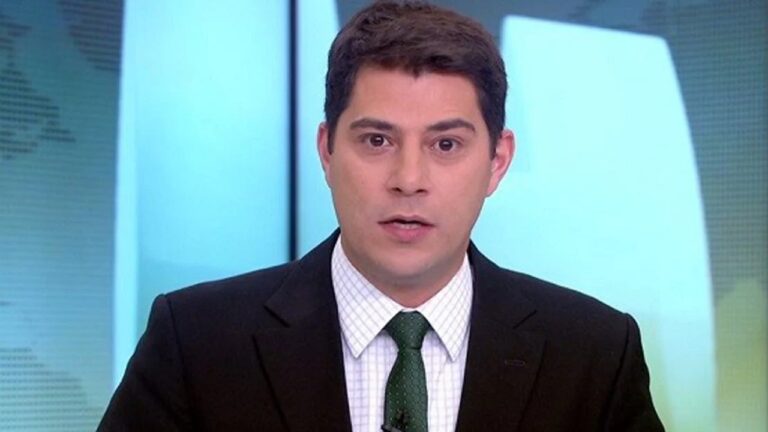Apresentador de telejornal usa terno escuro e gravata verde enquanto fala em estúdio com painel em tons de azul ao fundo.