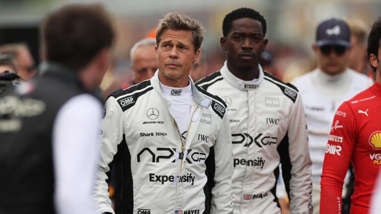 Dois homens vestindo macacões brancos de automobilismo caminham em área de paddock antes de corrida, cercados por integrantes de equipes; o uniforme exibe logotipos de patrocinadores e marcas automotivas.