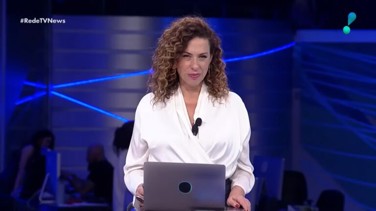 Fabíola Cidral, uma apresentadora de telejornal com cabelo cacheado e blusa branca, está em estúdio iluminado em tons de azul, atrás de um notebook, olhando diretamente para a câmera; no canto superior aparece a hashtag que indica a exibição do RedeTV! News.