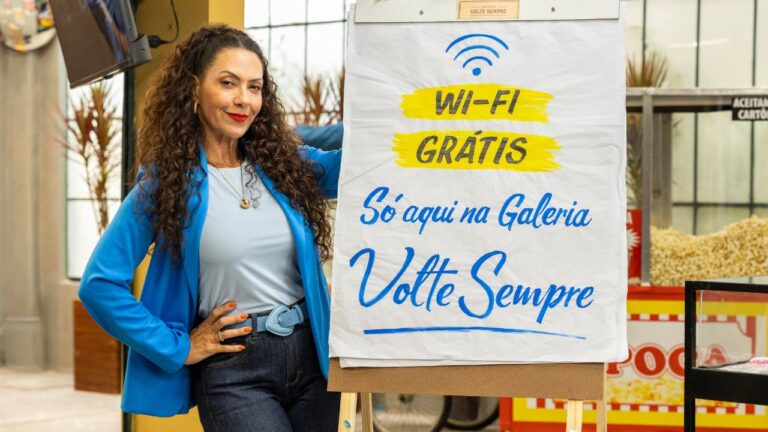 Fabiula Nascimento com cabelos cacheados e blazer azul posa ao lado de cartaz escrito "wi-fi frátis só aqui na galeria Volte Sempre" em cenário de loja