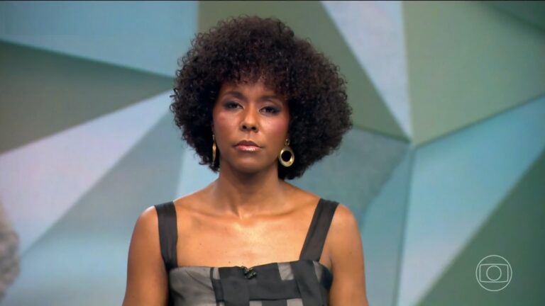 Maju Coutinho, que é uma mulher negra com cabelo crespo volumoso, veste roupa sem mangas em tons de preto e cinza e encara a câmera do Fantástico com expressão séria; ao fundo, painel geométrico em tons claros