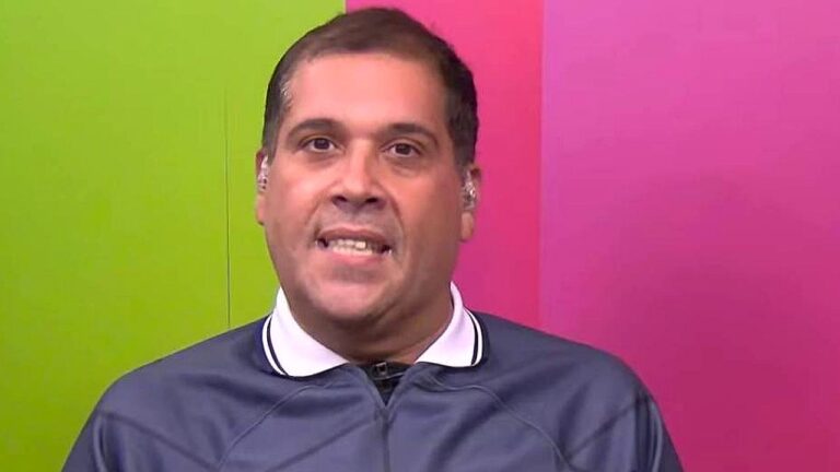 Homem aparece em estúdio de TV com fundo dividido em cores vibrantes, verde e rosa. Ele usa camiseta esportiva cinza com gola polo e fones de ouvido internos. A imagem é um close frontal, e ele parece estar falando diretamente para a câmera durante um programa ao vivo.