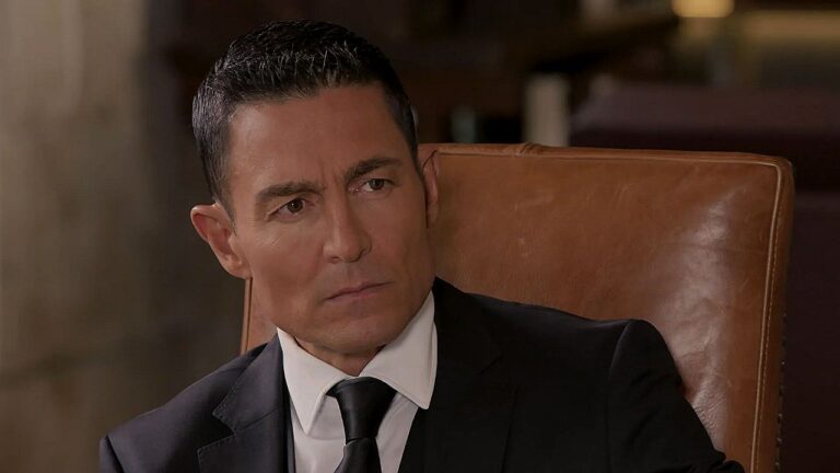 Homem de terno preto e gravata está sentado em poltrona de couro marrom, com expressão séria e olhar desconfiado voltado para alguém fora de cena. O ambiente interno elegante e iluminação suave sugerem cena dramática em contexto corporativo ou familiar.