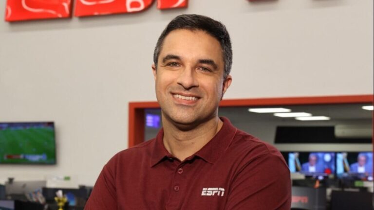 Apresentador com camisa bordô da ESPN sorri em redação esportiva com monitores exibindo transmissões ao fundo
