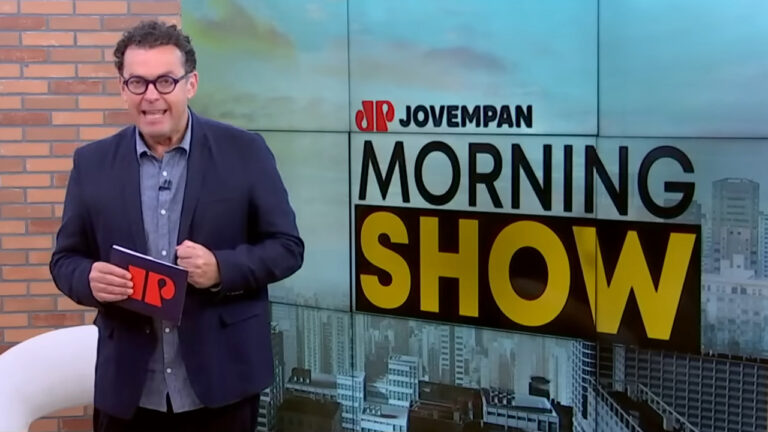 Fernando Rocha fala de óculos e blazer azul marinho segurando tablet da Jovem Pan em estúdio à frente do logotipo do programa Morning Show da Jovem Pan