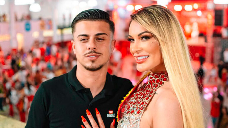 Flávio Giglioli de camisa polo preta ao lado de Andressa Urach, que usa vestido de pedrarias vermelhas, em evento com público ao fundo desfocado