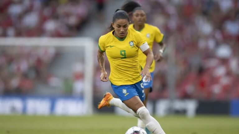 Jogadora de futebol em ação no gramado de um estádio, vestindo uniforme da seleção brasileira com camisa amarela de mangas curtas com detalhes verdes, número 9 em verde no centro do peito e escudo do Brasil à esquerda, short azul e meias brancas até o joelho, usando chuteiras laranja, cabelo escuro preso em rabo de cavalo alto, expressão concentrada e olhar voltado para a bola enquanto corre em velocidade, com o corpo levemente inclinado para a frente e a perna direita preparada para conduzir a bola branca com detalhes escuros, braço esquerdo com faixa de capitã branca, tatuagem visível no braço direito, outra jogadora da seleção ao fundo também de uniforme amarelo desfocada