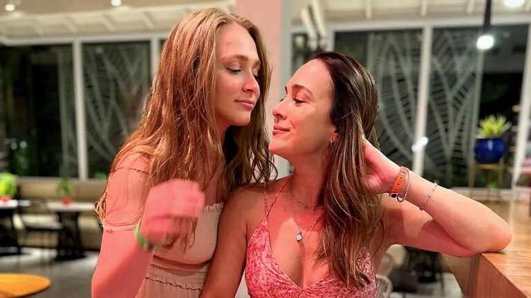 Duas mulheres estão próximas em ambiente interno bem iluminado, apoiadas em um balcão de madeira. Elas se olham com expressões suaves e sorrisos discretos, criando clima de intimidade e carinho. Ao fundo, há janelas amplas e mesas, sugerindo restaurante ou espaço social.