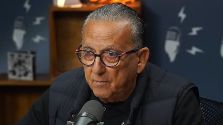 Galvão Bueno usa óculos redondos e colete preto em estúdio de podcast, com microfone à frente e ícones de som ao fundo em parede azul escura