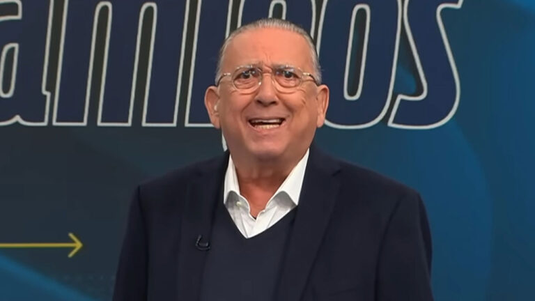 Galvão Bueno sorri de óculos e terno azul marinho sobre camisa branca em frente ao telão com logo do programa Galvão e Amigos em cenário de estúdio com fundo azul e detalhes amarelos