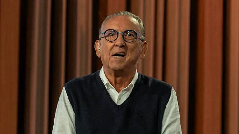 Homem idoso fala diante de cortinas marrons, usando óculos de armação redonda, camisa branca e colete escuro. Ele olha à frente com expressão engajada, em cenário que sugere gravação em estúdio ou participação em programa de televisão.