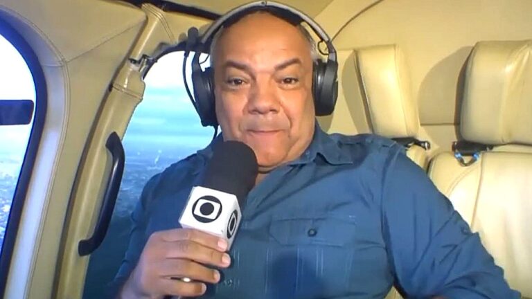 Homem com fones de ouvido fala ao microfone durante transmissão ao vivo dentro de uma aeronave. Ele segura microfone com logotipo de emissora de TV, sentado em poltrona clara, com janela ao fundo mostrando vista aérea.