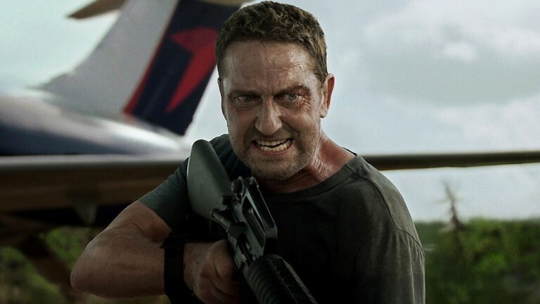 Um homem com expressão intensa e raivosa segura um rifle apontado para frente. Ele está suado e parece estar em uma situação de confronto. Ao fundo, há parte de um avião e uma área externa com vegetação, sugerindo uma cena de ação ao ar livre, possivelmente em um aeroporto ou pista de pouso.