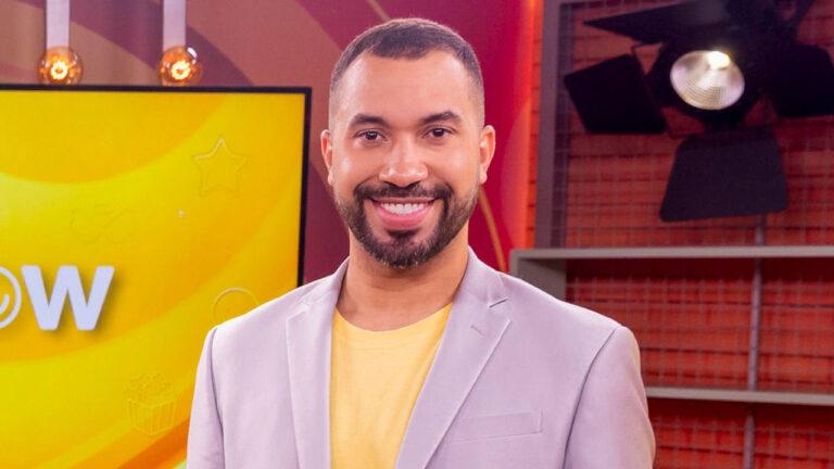 Gil do Vigor sorri de barba feita vestindo blazer cinza e camiseta amarela em estúdio de TV com painel vibrante ao fundo e equipamentos de iluminação