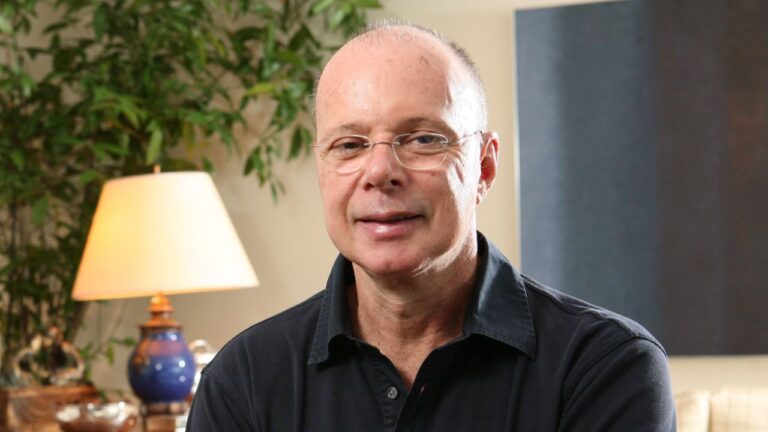 Autor e novelista Gilberto Braga sorri levemente usando óculos de grau e camisa polo escura em ambiente interno com luminária e planta ao fundo