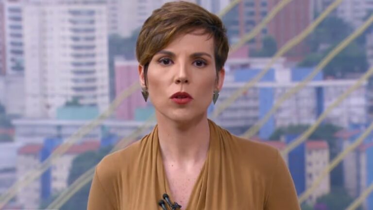 Jornalista com cabelo curto e blusa marrom apresenta telejornal em estúdio com vista de prédios ao fundo