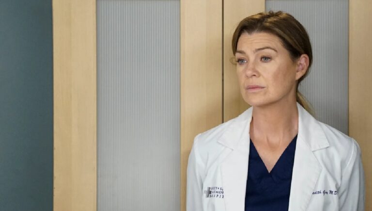 Mulher de meia-idade, vestindo jaleco branco sobre uniforme azul-marinho, aparece em ambiente hospitalar. Ela está diante de portas de madeira com painéis foscos e mantém expressão séria e contemplativa, olhando levemente para o lado. O crachá e a identificação no jaleco indicam que se trata de uma profissional de saúde em cena ambientada em hospital ou clínica.