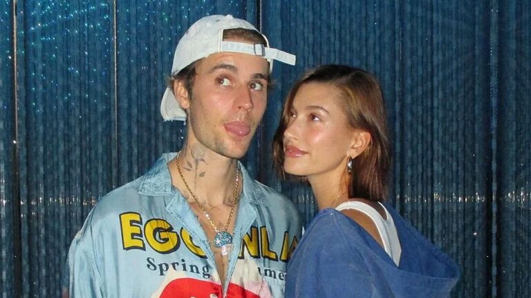 Casal posa diante de cortina azul com brilho. O homem usa boné claro virado para trás, camisa jeans estampada e colares, fazendo expressão divertida com a língua de fora. A mulher, com cabelo castanho na altura dos ombros, veste blusa azul e olha para ele com leve sorriso, sugerindo momento descontraído.