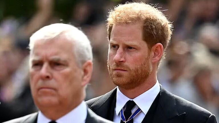 Dois homens vestindo ternos escuros aparecem em primeiro plano durante cerimônia pública ao ar livre. O homem à frente, ruivo e com barba, tem expressão séria e usa medalha pendurada no pescoço; atrás dele, um homem mais velho de cabelos brancos também mantém semblante solene. Ao fundo, uma multidão desfocada sugere evento oficial ou homenagem.