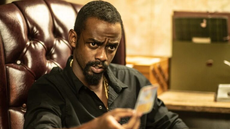 Homem negro sentado em cadeira de couro marrom escuro com encosto acolchoado observa com expressão tensa e concentrada uma carta que segura com a mão direita estendida à frente, usando camisa preta aberta no colarinho e corrente dourada no pescoço, com sobrancelhas franzidas, olhos fixos na carta e barba curta com cavanhaque, em ambiente interno com iluminação quente, mesa de madeira ao lado direito da imagem e arquivo ou caixa verde ao fundo sobre móvel, sugerindo escritório ou sala de interrogatório com clima sério e dramático