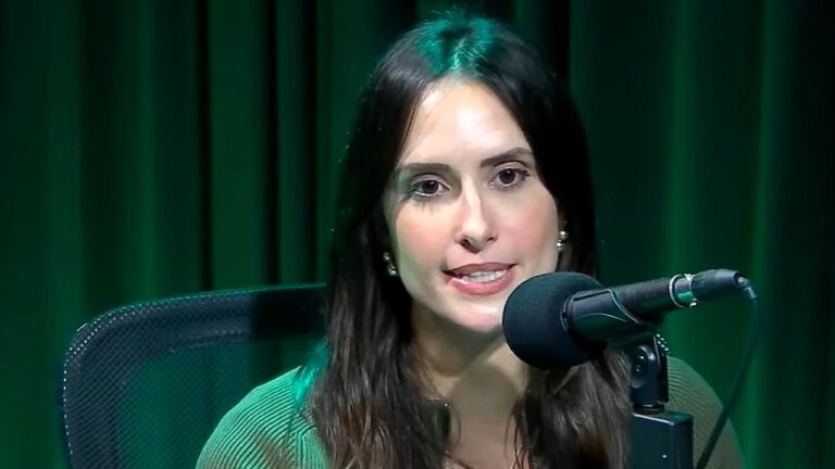 Mulher jovem está sentada diante de um microfone profissional, como em um estúdio de rádio ou podcast. Ela tem cabelo longo e escuro, usa brincos discretos e veste uma blusa verde. Sua expressão é séria e focada, como se estivesse falando ou respondendo a uma pergunta. Ao fundo, há cortinas ou painéis verdes e iluminação que reforça o tom do ambiente, sugerindo uma gravação em estúdio.