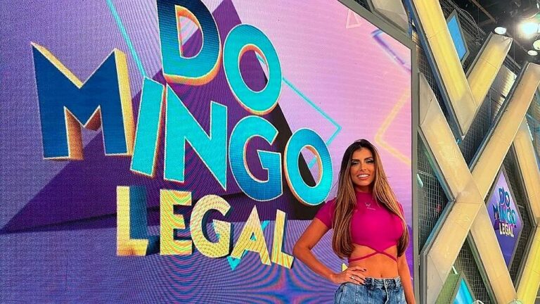 Apresentadora posa sorridente em estúdio ao lado de um grande painel colorido com a frase “Domingo Legal” em destaque. Ela veste top rosa e jeans, com uma das mãos na cintura, enquanto a estrutura moderna do cenário aparece ao fundo, sugerindo gravação de programa de televisão.