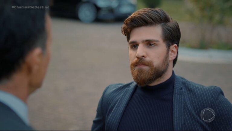 Iskender, um homem jovem de barba e cabelo castanho bem penteado, veste blazer escuro listrado e blusa de gola alta preta, olhando com expressão séria para outra pessoa em cena na novela Chamas do Destino; ao fundo, ambiente externo com rua desfocada