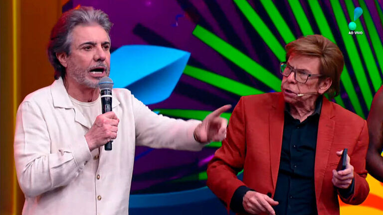 João Kleber segura microfone e aponta para o lado, enquanto Nelson Rubens, de paletó vermelho e óculos, observa em estúdio colorido da RedeTV
