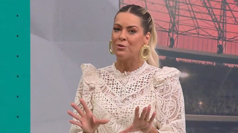 Renata Fan, uma mulher de cabelos loiros presos em rabo de cavalo, fala no estúdio do Jogo Aberto, vestindo blusa branca rendada com detalhes vazados e brincos grandes dourados, gesticulando com as mãos enquanto explica algo.