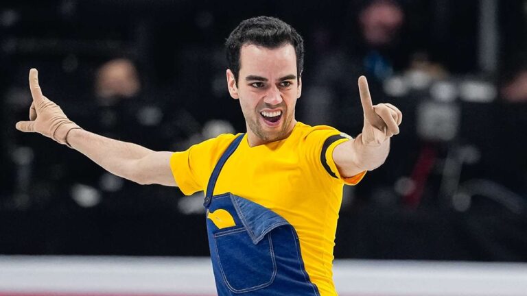 Patinador artístico com fantasia amarela e azul faz performance sobre o gelo com expressão de entusiasmo